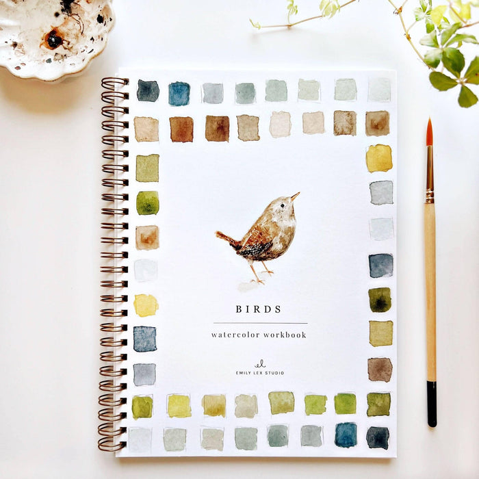 A spiral-bound watercolor workbook titled 'BIRDS' by Emily Lee Studio, featuring a watercolor bird illustration in the center and a border of watercolor color swatches. The workbook is displayed on a white surface with a paintbrush, a small dish, and green leaves.