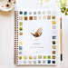A spiral-bound watercolor workbook titled 'BIRDS' by Emily Lee Studio, featuring a watercolor bird illustration in the center and a border of watercolor color swatches. The workbook is displayed on a white surface with a paintbrush, a small dish, and green leaves.