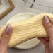 A close-up of a person's hands with manicured nails stretching a pale yellow, textured squishy toy over a white plate.