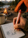 A hand holds a fishing-themed pen with a red and yellow striped body and white line details, writing in a spiral notebook labeled 'CAMP NOTES'. A rustic mug sits nearby, with a campfire, mountains, and an orange tent in the background.