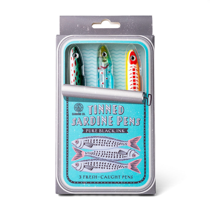 A packaged set of three novelty tinned sardine pens in a clear plastic blister pack with a gray backing card. The pens are decorated like sardines with green, blue, and red-and-white striping, and the package text reads 'Tinned Sardine Pens' and 'Pure Black Ink'.