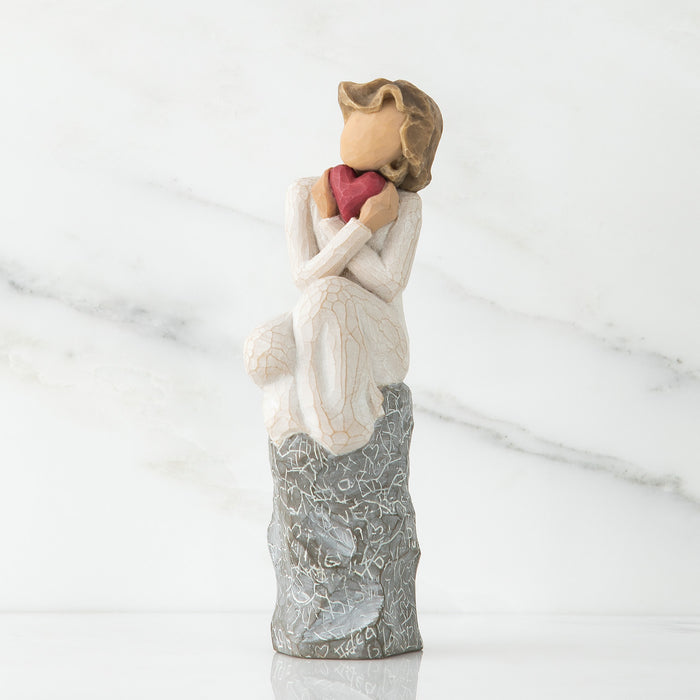 Figure in cream dress, seated on a gray rock, with knees drawn up to chest, holding red heart up to face. Rock etched with words and symbols of love