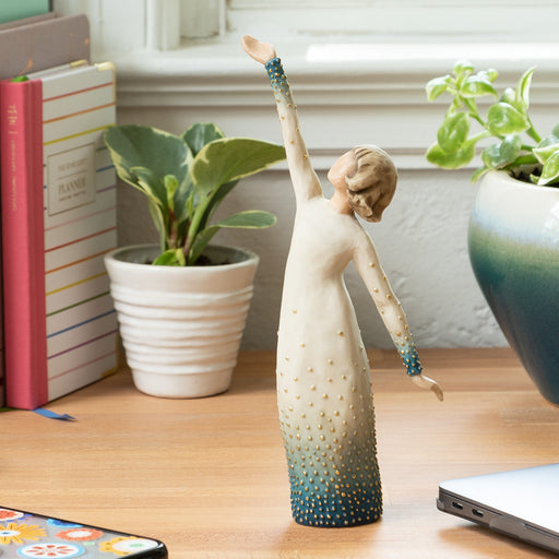 Decorative figurine of a woman in a yoga pose on a desk with plants and books in the background.