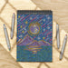 Decorative notebook with celestial design on a wooden surface with dot markers.