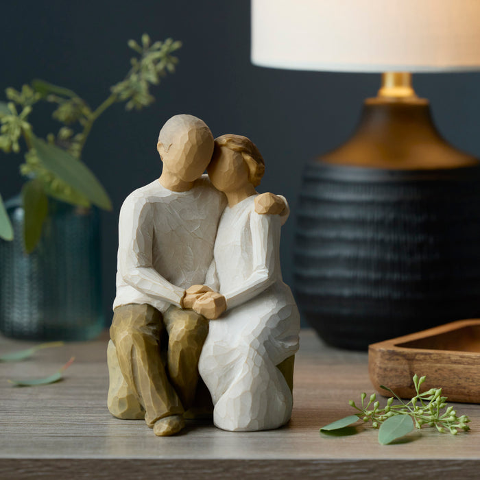 Wooden figurine of a couple embracing on a wooden surface with a lamp and plant in the background.