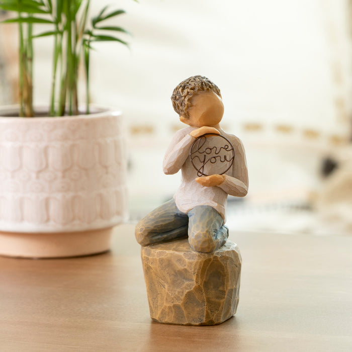 Decorative figurine of a child holding a heart with 'love you' on a wooden base, placed on a surface with a blurred background.