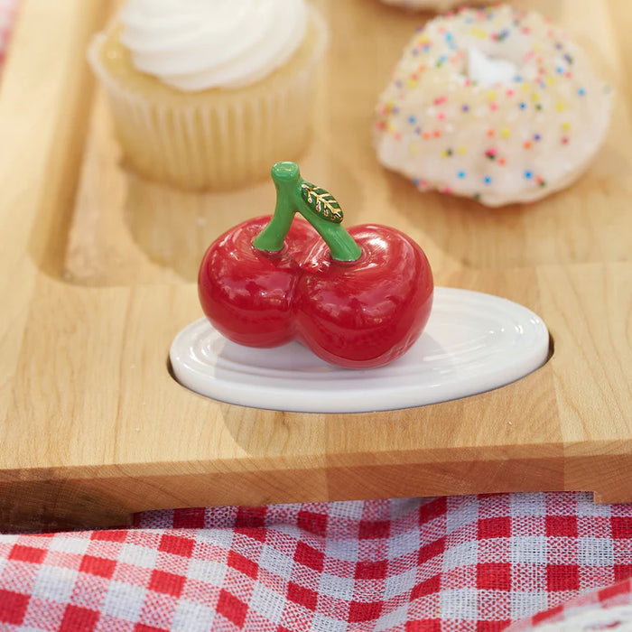 Decorative cherry on a white base with a wooden surface and pastries in the background.