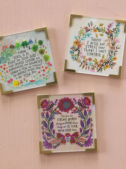Three square decorative coasters with gold-toned metal corner protectors, featuring colorful floral illustrations and inspirational quotes on a light pink background.