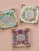 Three square decorative coasters with gold-toned metal corner protectors, featuring colorful floral illustrations and inspirational quotes on a light pink background.