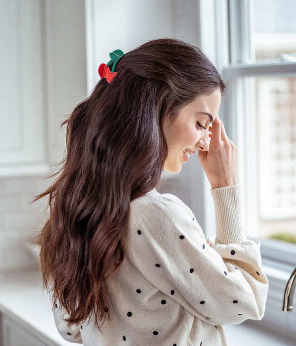 Woman with a decorative hair clip in her hair, wearing a polka dot sweater, standing by a window.