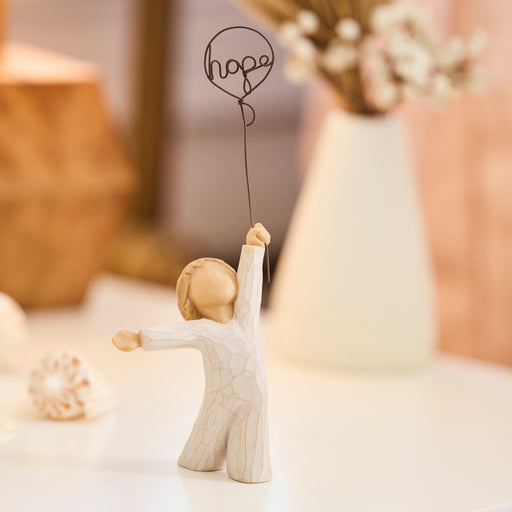 Standing figure in cream onesie, holding wire balloon with the word 'hope' written in wire inside it