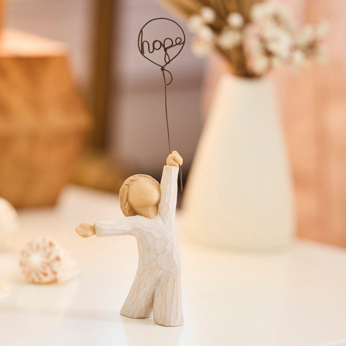 Standing figure in cream onesie, holding wire balloon with the word 'hope' written in wire inside it