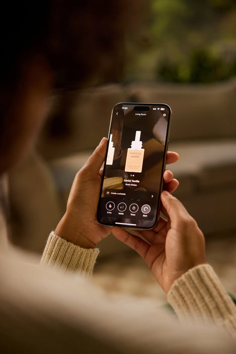 A person holding a smartphone outdoors, showing a beauty app screen with a skincare bottle on the display. The person is wearing a beige ribbed sweater, and the background is softly blurred.