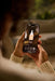 A person holding a smartphone outdoors, showing a beauty app screen with a skincare bottle on the display. The person is wearing a beige ribbed sweater, and the background is softly blurred.