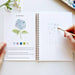 Person coloring a hydrangea flower in a coloring book with a pen.