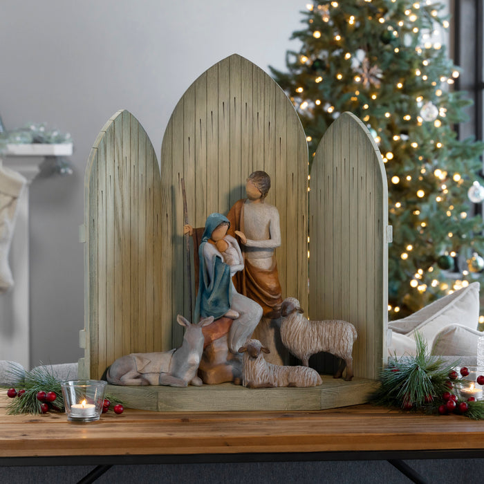 A wooden nativity scene display with three arched panels and a central figurine of Mary and Joseph with baby Jesus and sheep, styled on a wooden table with a lit Christmas tree in the background.