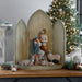 A wooden nativity scene display with three arched panels and a central figurine of Mary and Joseph with baby Jesus and sheep, styled on a wooden table with a lit Christmas tree in the background.