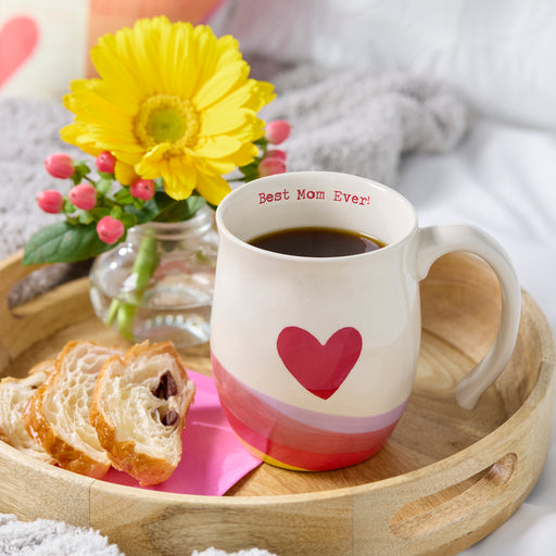 Ceramic mug with 'Best Mom Ever!' text and heart design on a wooden tray with bread and flowers.