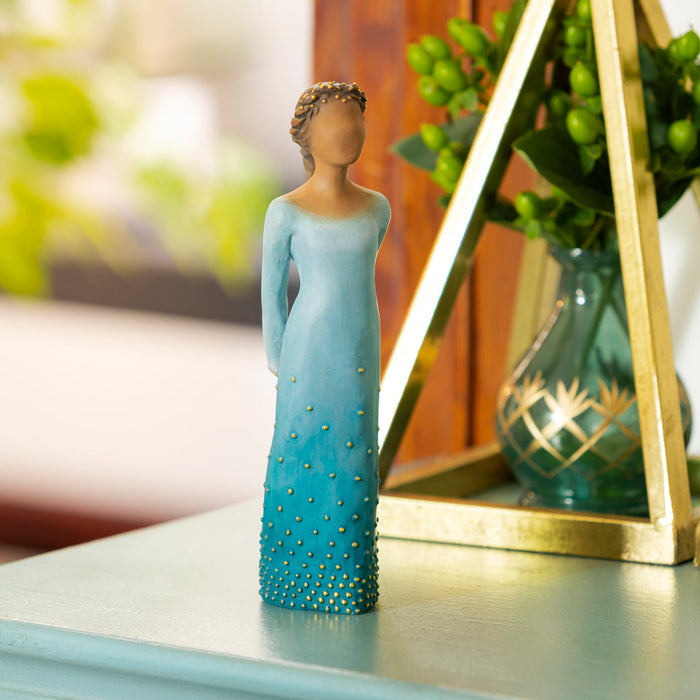 Standing figure with arms clasped behind back, hair and deep blue ombré dress are dotted with gold leaf. Figure has darker skin and hair color
