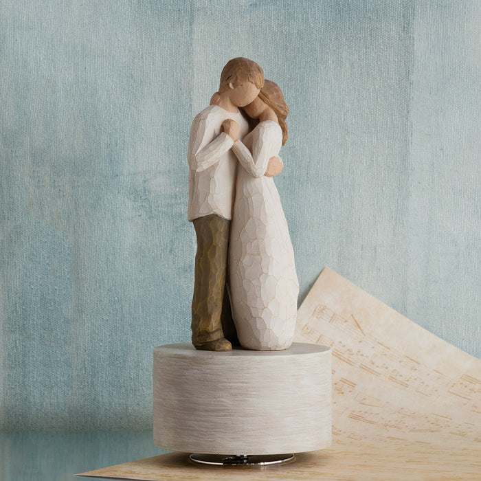 Statue of a couple embracing on a wooden base against a textured blue wall.
