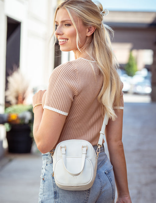 Woman wearing a beige sweater and light blue jeans with a white crossbody bag outdoors.