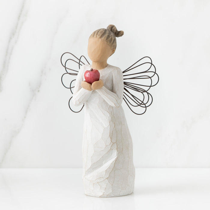 Standing angel in cream dress with wire wings, holding large, shiny red apple in outstretched hands