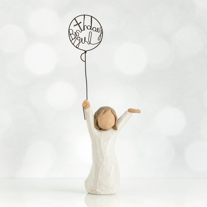 Standing figure in cream dress, holding wire ballon with the words 'birthday girl' written in wire inside it