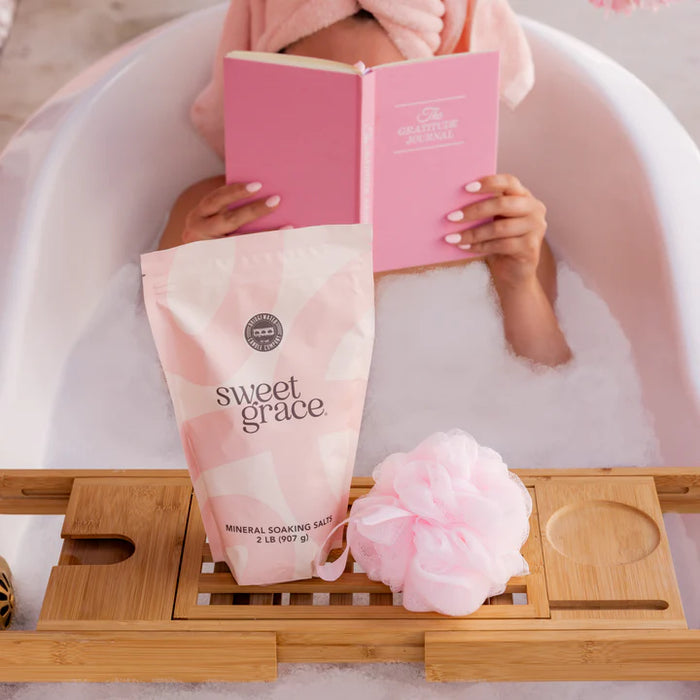 Person in a bathtub with a pink book and 'sweet grace' product, surrounded by bubbles and a bath tray.